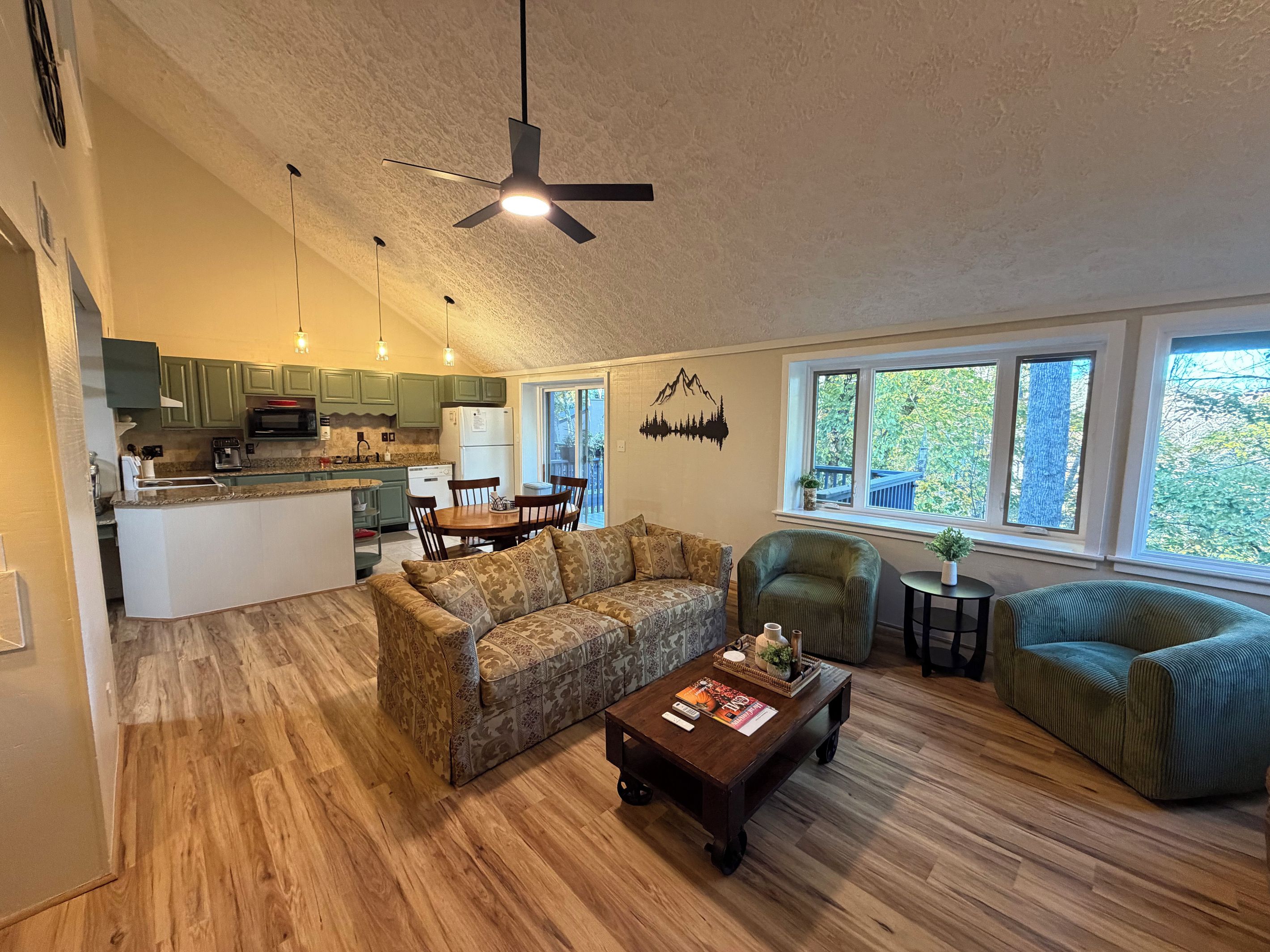 A spacious living area with a sofa, coffee table, and a dining table with chairs. A kitchen with green cabinets and a microwave is visible in the background.
