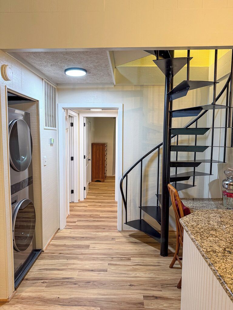 Un pasillo con suelo de madera, una lavadora, una escalera con barandilla de metal y una puerta que da a otra habitación.