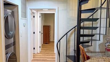 Un pasillo con suelo de madera, una lavadora, una escalera con barandilla de metal y una puerta que da a otra habitación.