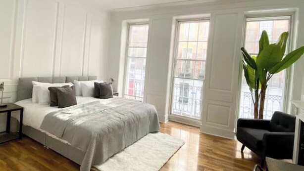 Ein modernes Schlafzimmer mit einem großen Bett, zwei Fenstern, einer Pflanze und einem Sessel.