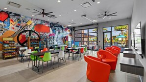 Un café animé, avec des chaises aux couleurs vives, un mur décoré d’une fresque et une vue sur l’espace extérieur de la piscine.