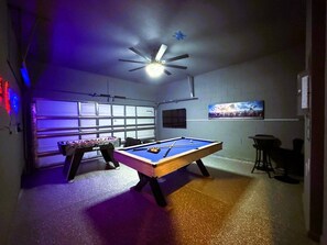 Una mesa de billar junto a una mesa de fútbol de mesa en una habitación con ventilador de techo, ventana y televisor en la pared.