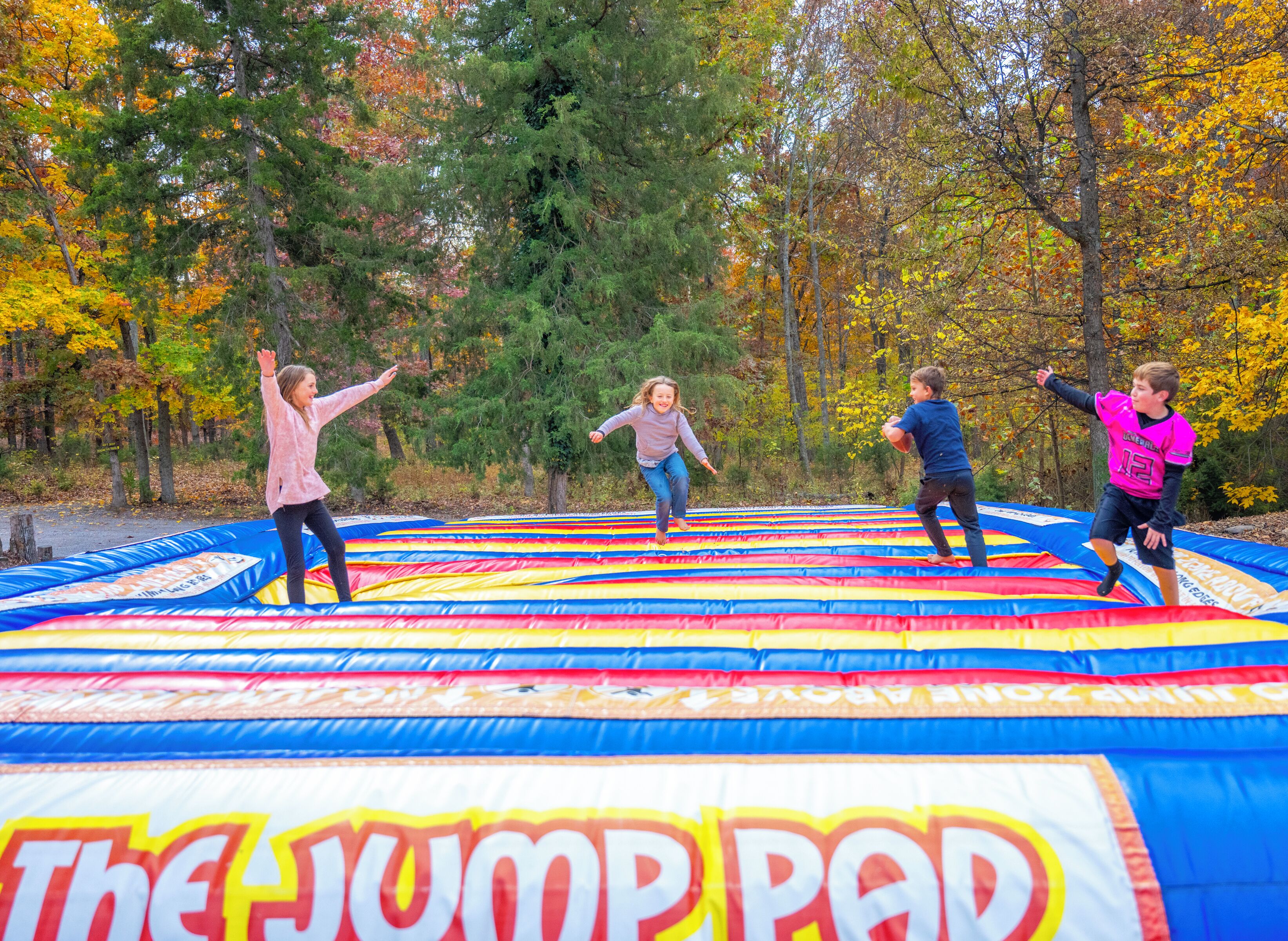 Ryhmä ihmisiä hyppii värikkäällä, ilmatäytteisellä alustalla, jossa on teksti ”The Jump Pad”, metsäisellä alueella.