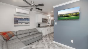 Una sala de estar moderna con un sofá seccional gris, un ventilador de techo, un televisor de pantalla plana montado en la pared y una cocina al fondo.