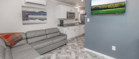 Un salon moderne avec un canapé d’angle gris, un ventilateur de plafond, un téléviseur à écran plat fixé au mur et, au fond, un coin cuisine.