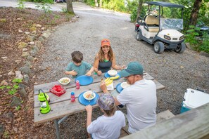 A group of people dining at an outdoor table with a picnic setup, including food and drinks, in a natural setting with a golf cart in the background. - Endless Caverns RV Resort & Cavern Tours (New Market)