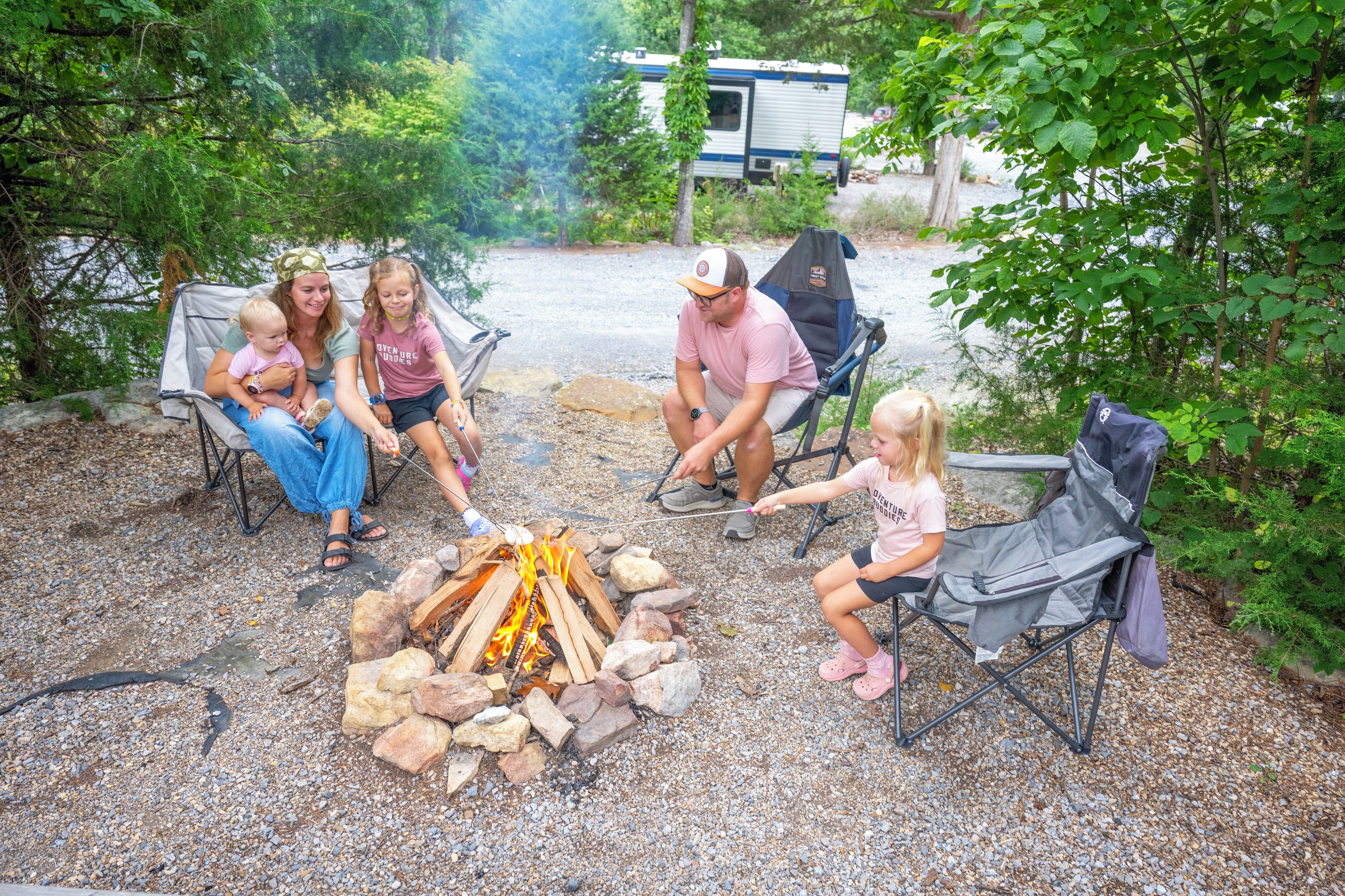 A group of people around a campfire in a wooded area with a trailer in the background.