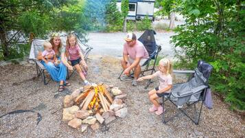 A group of people around a campfire in a wooded area with a trailer in the background.