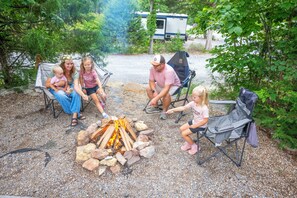 A group of people around a campfire in a wooded area with a trailer in the background. - Endless Caverns RV Resort & Cavern Tours (New Market)