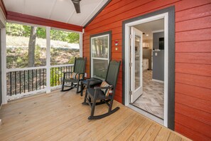 Une véranda couverte avec un plancher en bois, deux fauteuils à bascule et une porte donnant sur un coin cuisine.