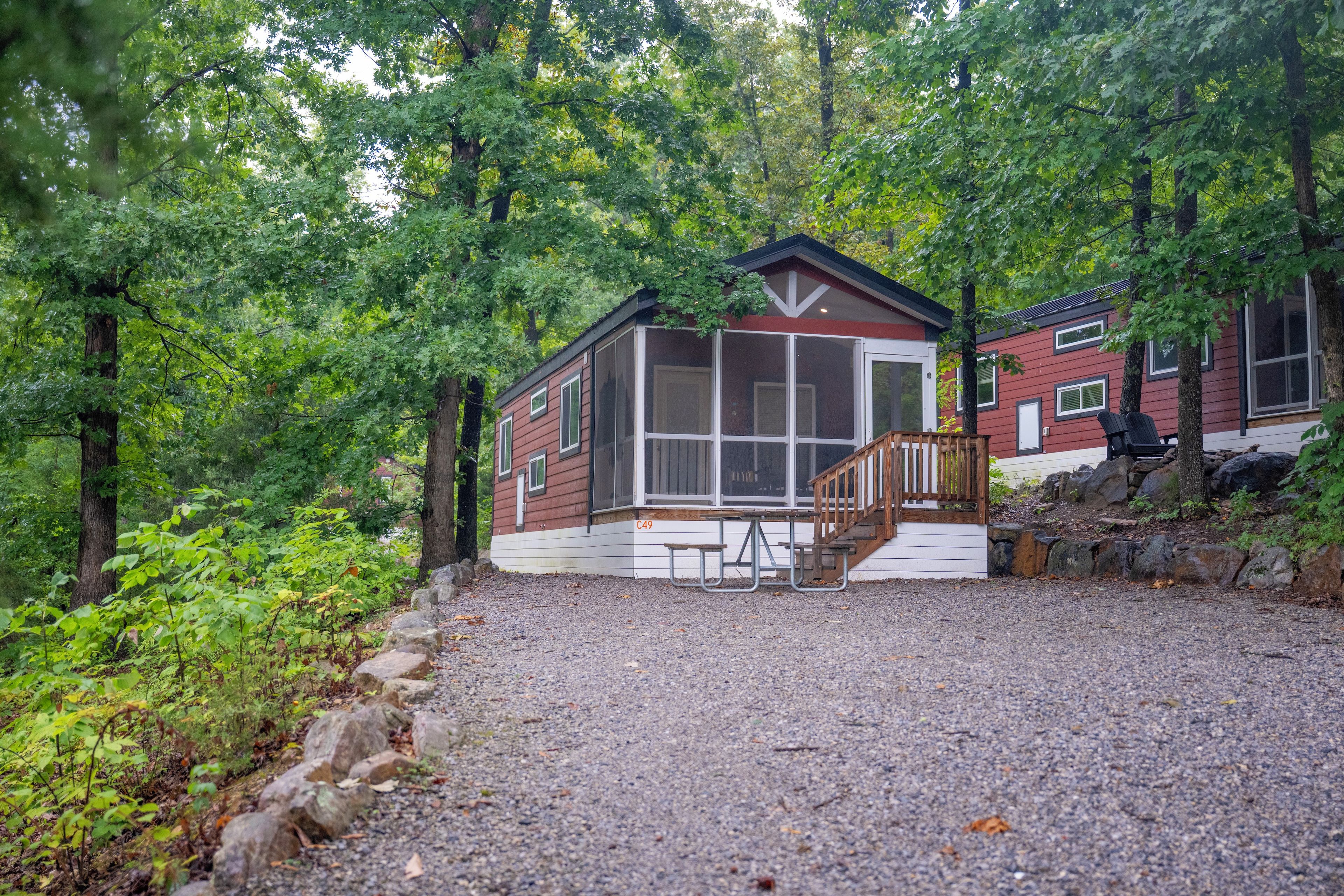 A cabin with a covered porch, a gravel driveway, and a forested backdrop.