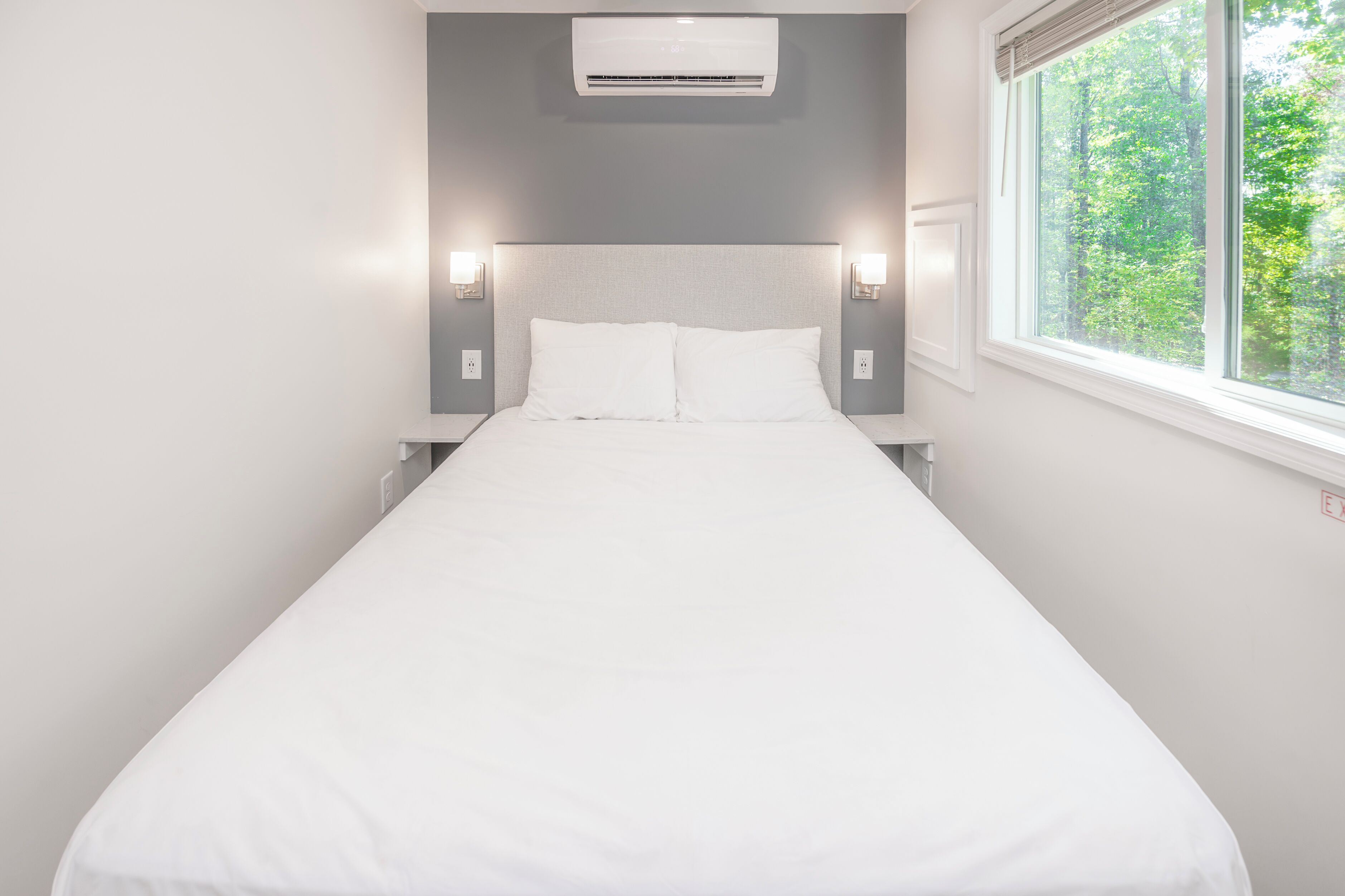Minimalistinen hotellihuone, jossa on yksi sänky, sen yläpuolella ilmastointilaite ja ikkunasta avautuu näkymä puihin.