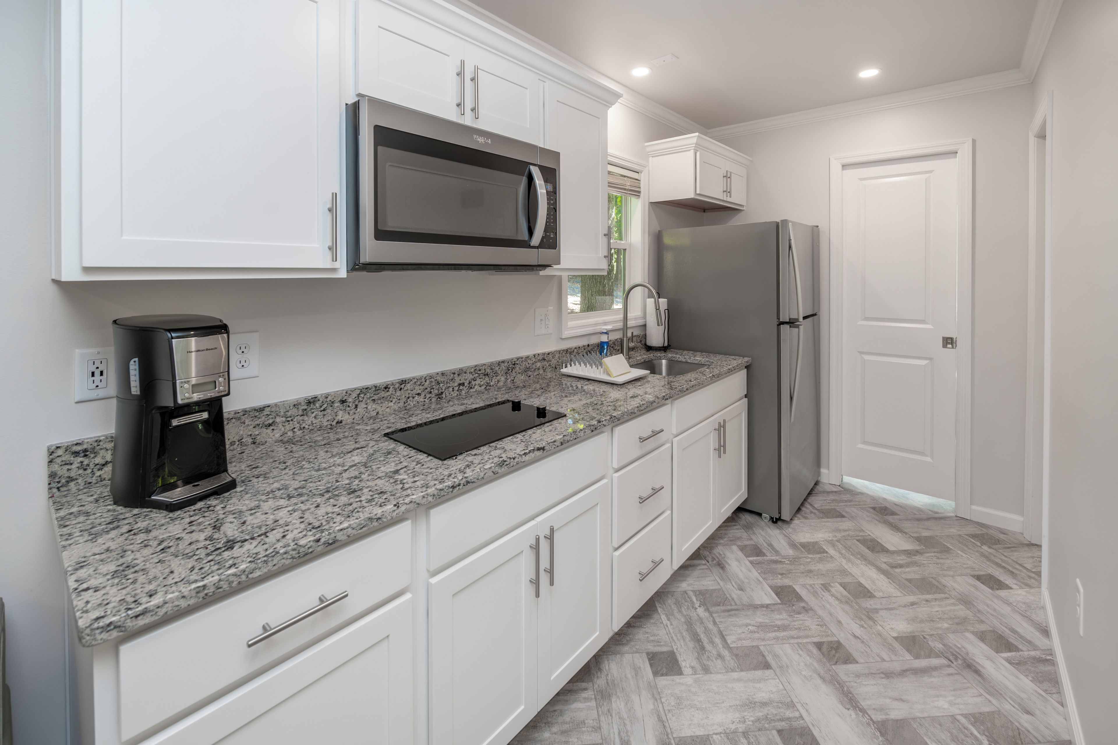 A modern kitchen with white cabinets, a granite countertop, a built-in microwave, a coffee maker, a stainless steel refrigerator, and a herringbone-patterned floor.