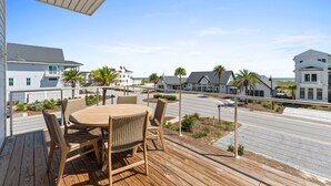 Een houten terras met een ronde tafel en stoelen, uitkijkend op een straat met huizen en een heldere lucht.