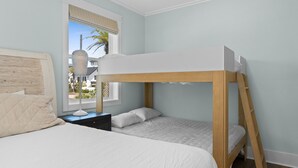 Een stapelbed met houten frame en witte beddengoed, een nachtkastje met een lamp, en een raam met uitzicht op palmbomen.