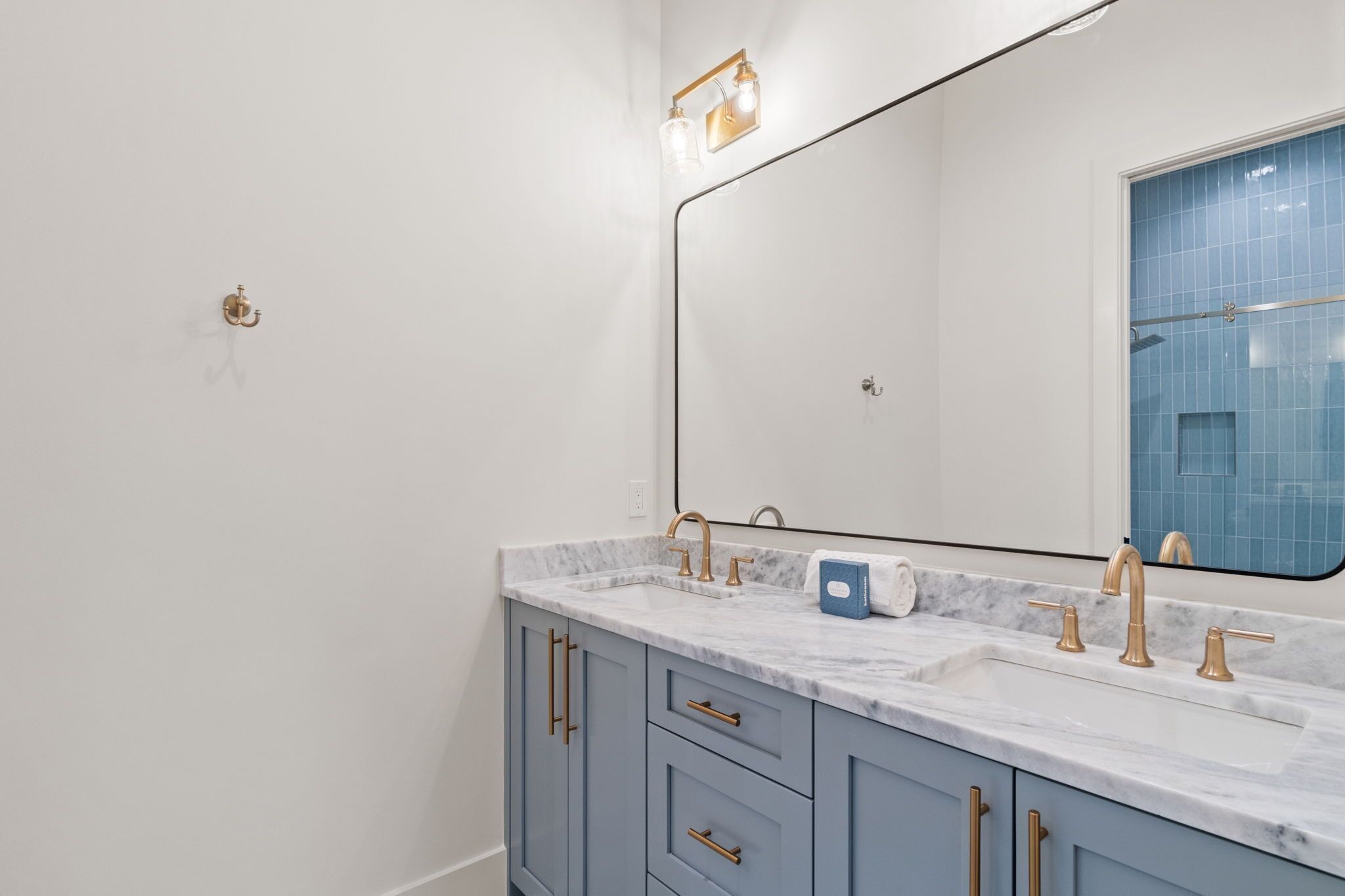 Une salle de bain dotée d’un double lavabo, de robinets dorés, d’un comptoir en marbre et d’un grand miroir, avec une cabine de douche aux carreaux bleus.