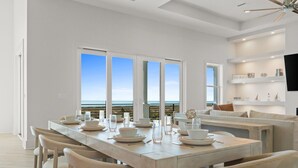 A modern dining area with a long table set for six, large windows with a view of the ocean, and a cozy seating area with a TV on the wall. - Ocean Breeze: Unobstructed Views, Private Elevated Pool, Pet Friendly, Sleeps 19 (Mexico Beach)