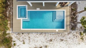 Ein Luftbild eines rechteckigen Schwimmbeckens mit umlaufender Holzterrasse, vor einer sandigen Küste mit vereinzelter Vegetation.