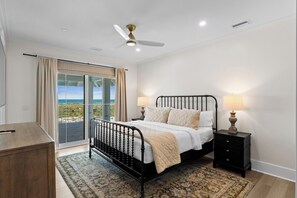 Ein Schlafzimmer mit einem Bett, Nachttischlampen, einem Nachttisch, einer hölzernen Kommode, einem Balkon mit Vorhängen und einem Deckenventilator.