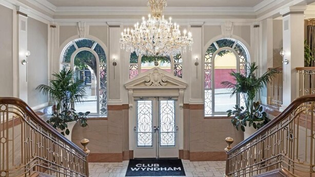Une entrée imposante avec un lustre, une porte à double battant et un paillasson portant l'inscription « CLUB WYNDHAM ».
