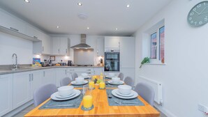 A modern kitchen with white cabinets, a central island, and a dining area set for six with a wooden table. - Triple Driveway | Telford | Central | Long Stays (Telford)