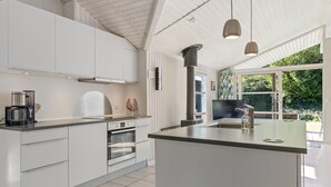 Een moderne keuken met witte kasten, een groot eiland, roestvrijstalen apparatuur, hangende lampen en uitzicht op groen.