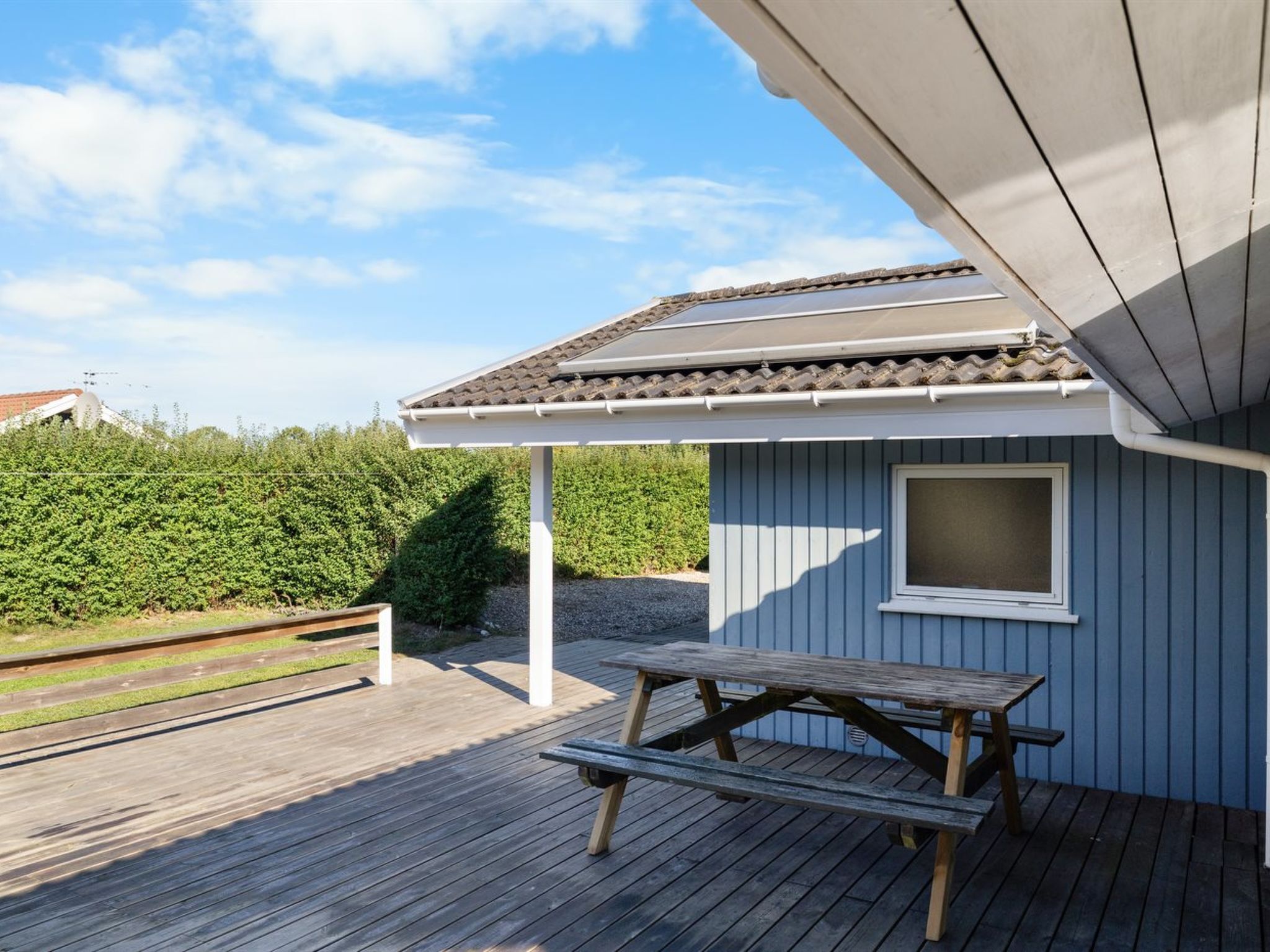 A covered outdoor area with a wooden bench and a table, a window, and a tiled roof.