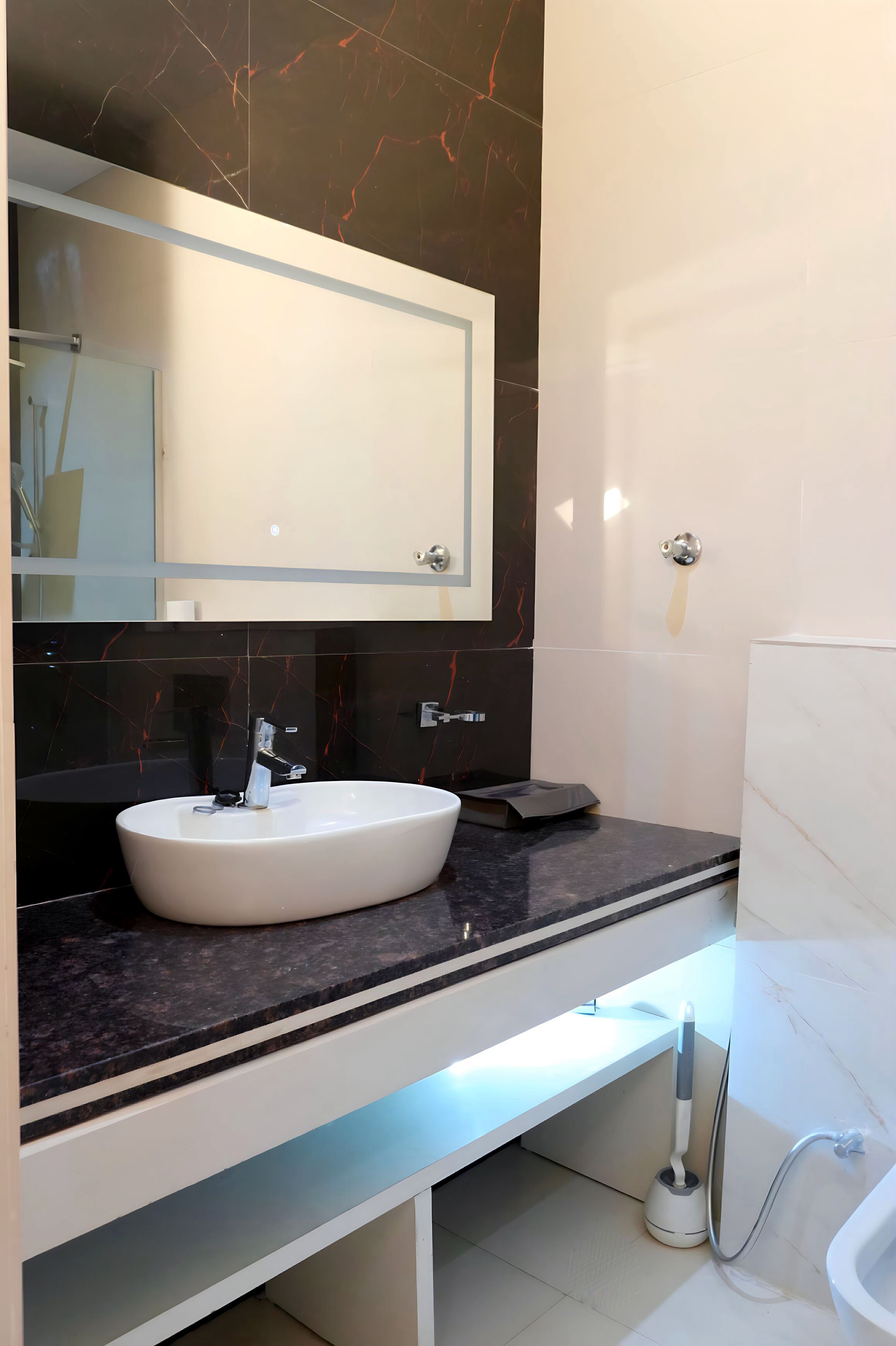 Une salle de bain moderne avec un grand miroir, un lavabo blanc, un plan de toilette en marbre foncé et une douchette à main.