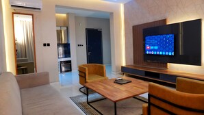 A modern living room with a wooden coffee table, two armchairs, a flat-screen TV, and a kitchen in the background. - 2020 Apartment Guzape (Abuja)