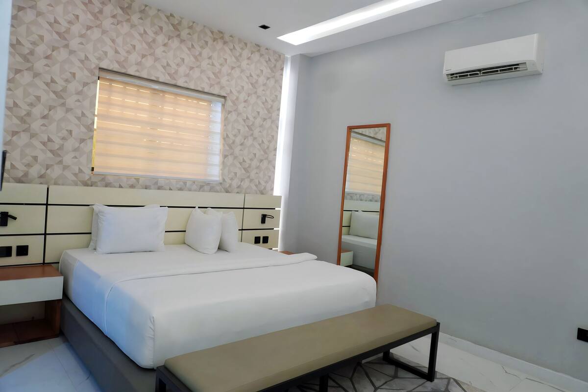 Une chambre d’hôtel moderne avec un grand lit, une table de chevet, un banc, une fenêtre avec des stores, un climatiseur fixé au mur et un miroir.