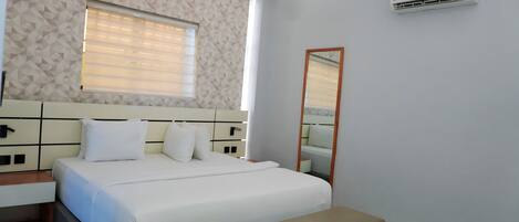 A modern hotel room with a large bed, a bedside table, a bench, a window with blinds, a wall-mounted air conditioner, and a mirror.