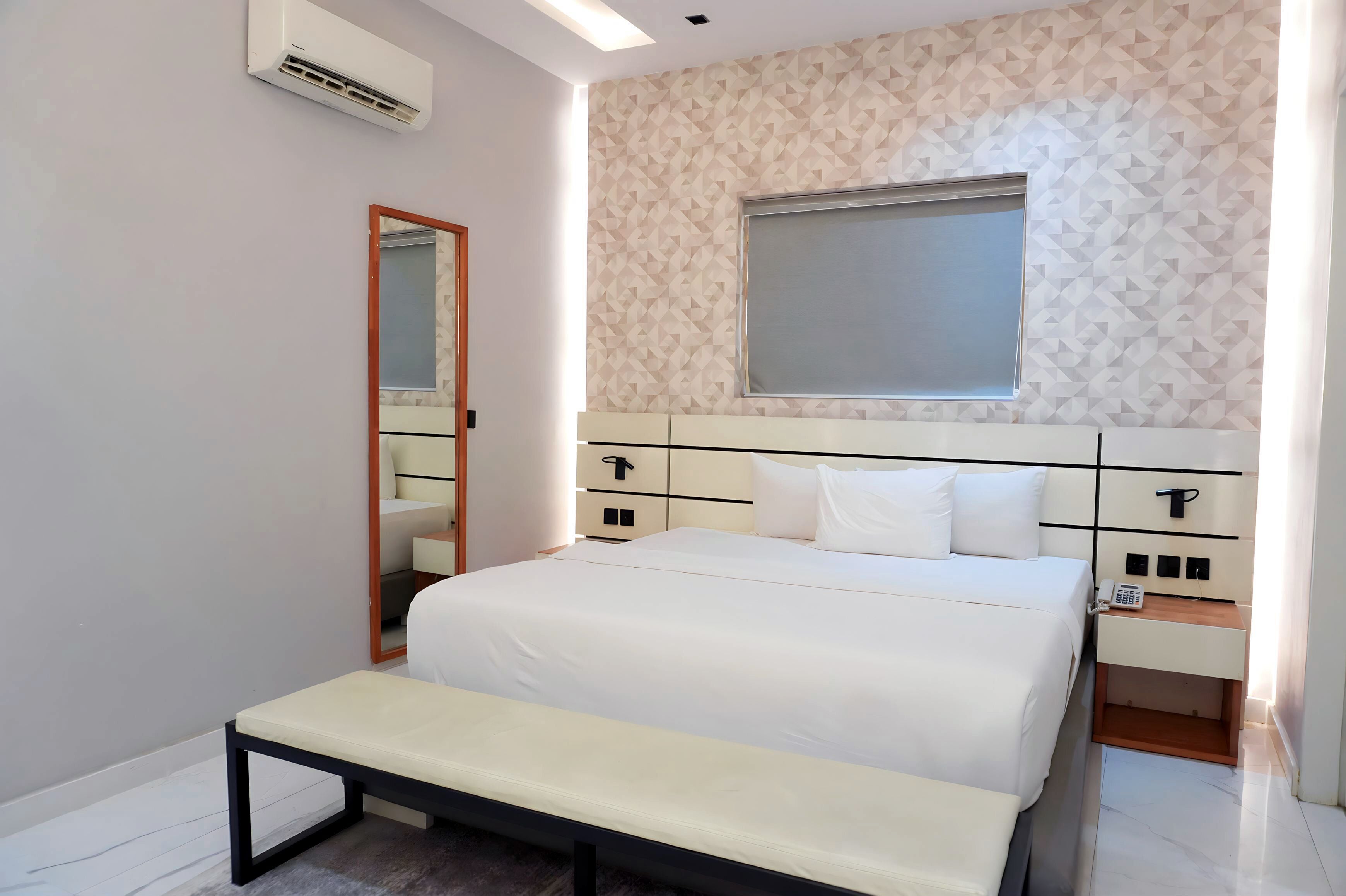 A modern bedroom with a large bed, bedside tables, a bench, a mirror, and a window with a geometric wall pattern.