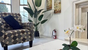 Un salón con un sillón estampado, una planta grande en maceta, un florero con flores blancas y un extintor en la pared.