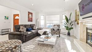 Un salon spacieux avec un canapé gris, un pouf à motifs et une table basse blanche. On y trouve également une grande plante, une cheminée et un téléviseur qui diffuse l’émission « Hospik ».
