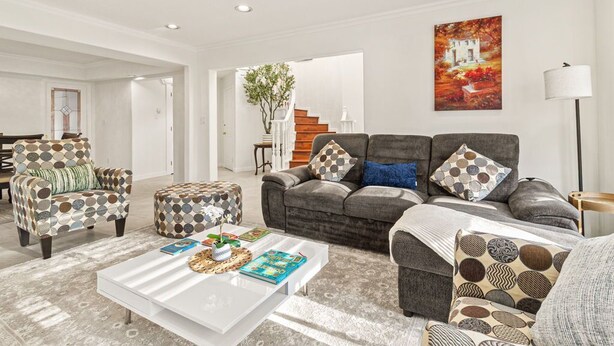 Una sala moderna con un sofá gris, un sillón estampado y un reposapiés, una mesa de centro blanca con libros y un objeto decorativo, y una escalera al fondo.