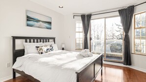 Un dormitorio con una cama de madera, ropa de cama blanca, un cuadro en la pared y ventanas grandes con cortinas.