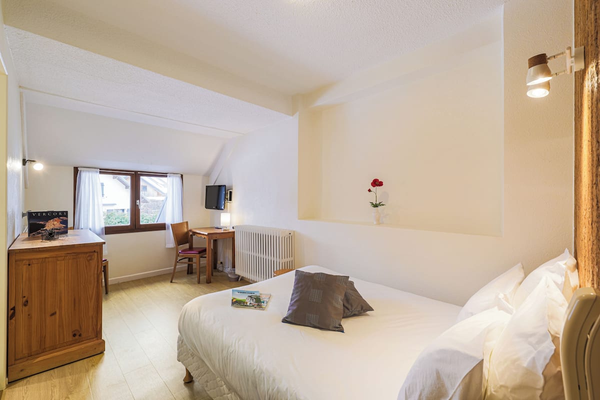 Un dormitorio con cama, un armario de madera, un escritorio, una silla, un radiador, un televisor y una ventana con cortinas.