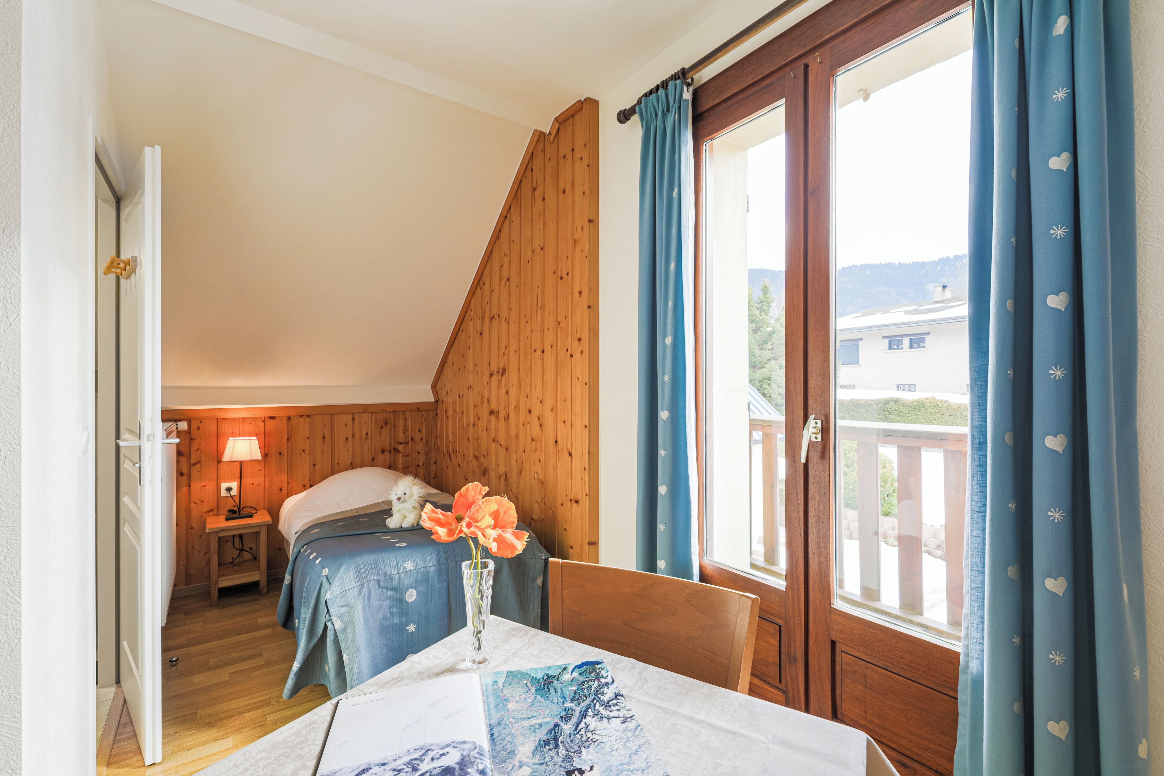 A bedroom with a wooden sloped ceiling, a bed with a blue bedspread, a nightstand with a lamp, and a window with blue curtains.