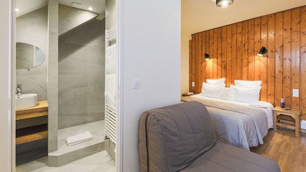 Ein modernes Badezimmer mit Dusche, Waschbecken und Spiegel. Im angrenzenden Raum befindet sich ein Bett mit Kissen an einer Holzwand.
