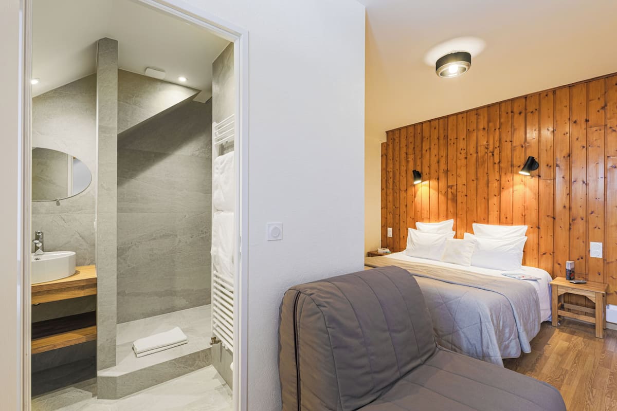 Un baño moderno con ducha, lavabo y espejo. En la habitación contigua, hay una pared de madera con una cama y almohadas.