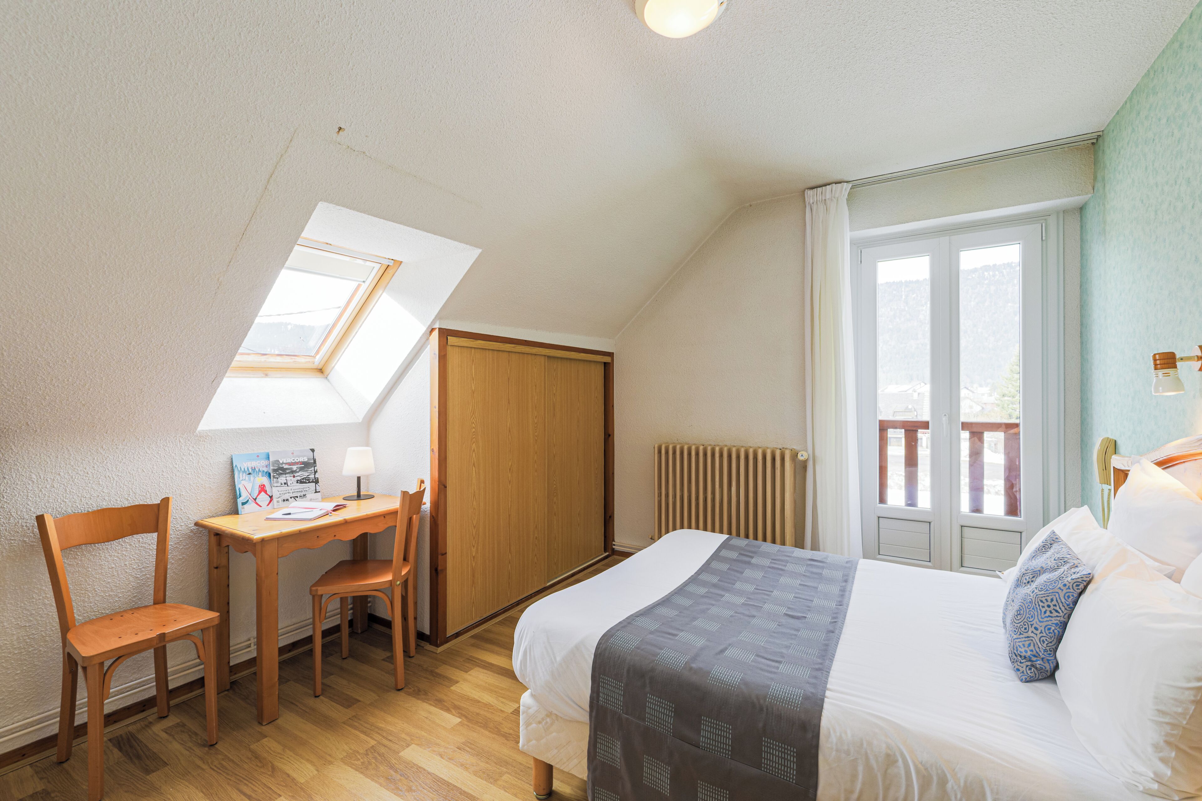 Une chambre avec un puits de lumière, un bureau en bois, une chaise, un lit avec des oreillers et une fenêtre donnant sur un balcon.