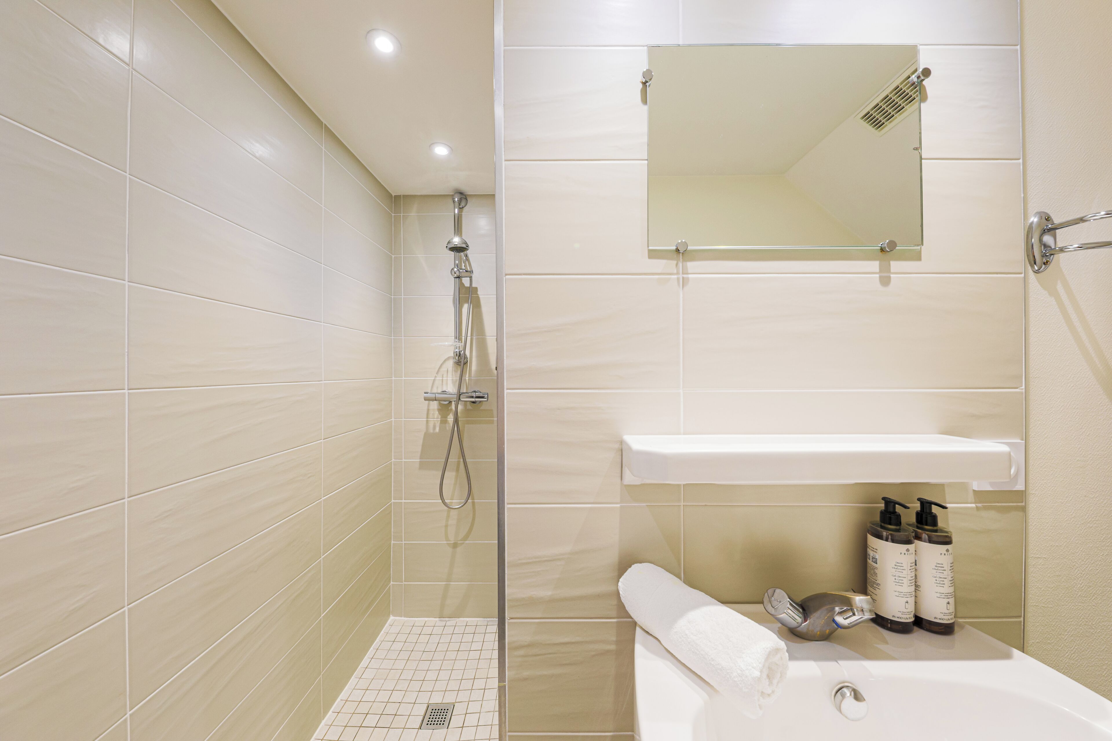 A bathroom with beige tiles, a shower, a sink with two bottles of shampoo, and a towel.