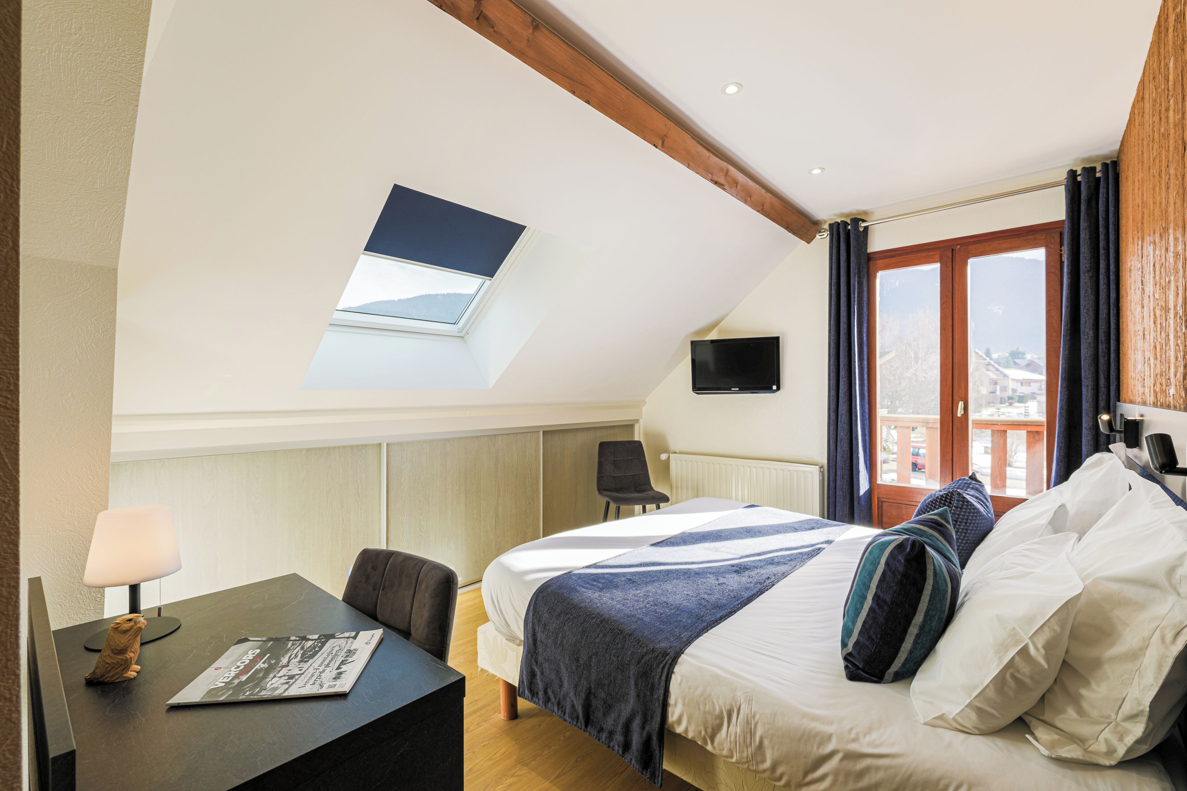 A bedroom with a skylight, a bed with pillows, a desk with a lamp, and a TV on the wall.