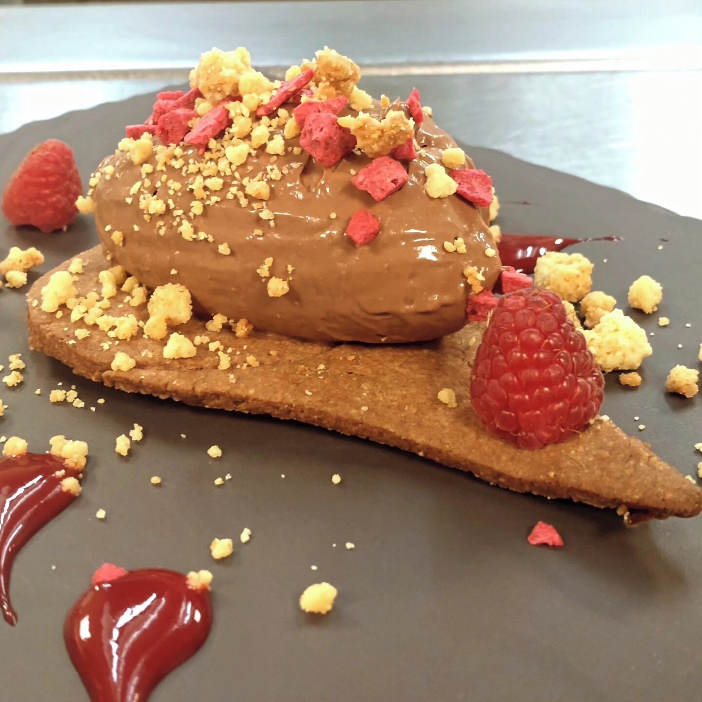 A dessert on a plate with a chocolate coating, crumbled topping, and raspberries.