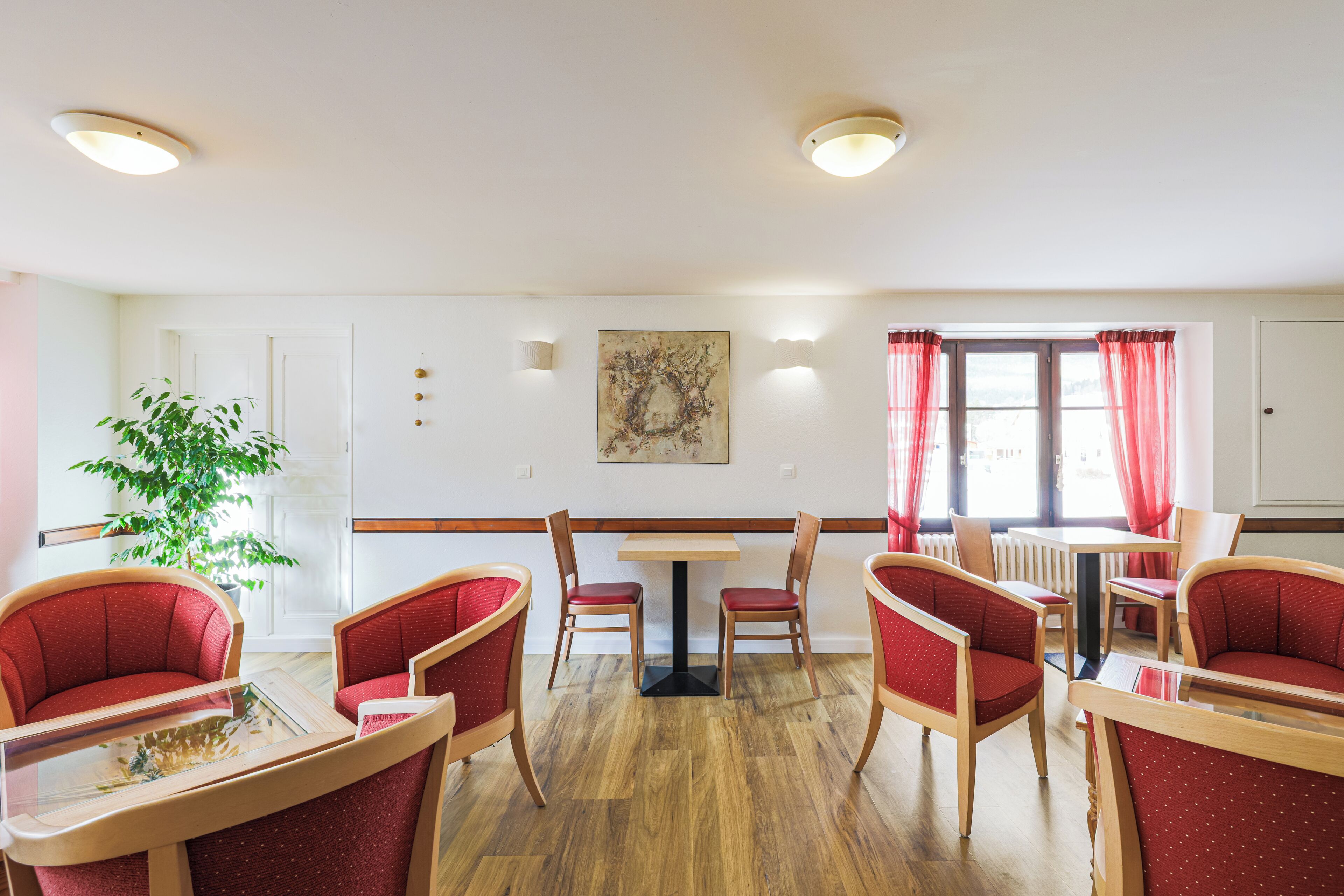 A dining area with wooden tables and red upholstered chairs, a potted plant, and a painting on the wall.