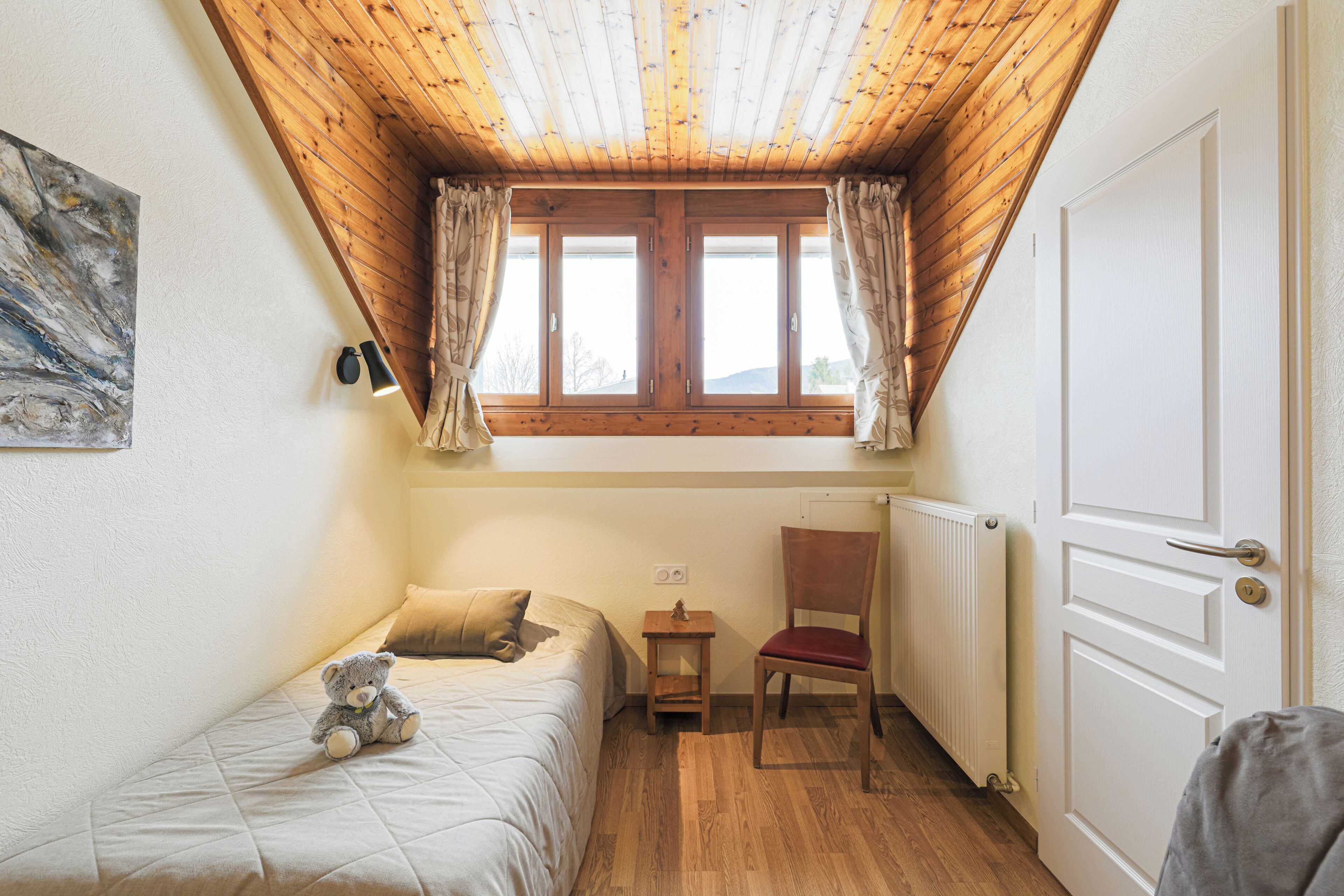 A bedroom with a wooden ceiling, a window with curtains, a bed with a teddy bear, a chair, and a radiator.