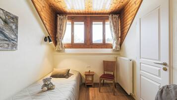 A bedroom with a wooden ceiling, a window with curtains, a bed with a teddy bear, a chair, and a radiator.