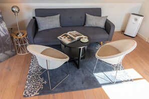 Ein modernes Wohnzimmer mit einem grauen Sofa, zwei Korbstühlen, einem kleinen runden Tisch mit einer Zeitschrift und einer Tasse, einer Stehlampe und einem weißen Heizkörper.