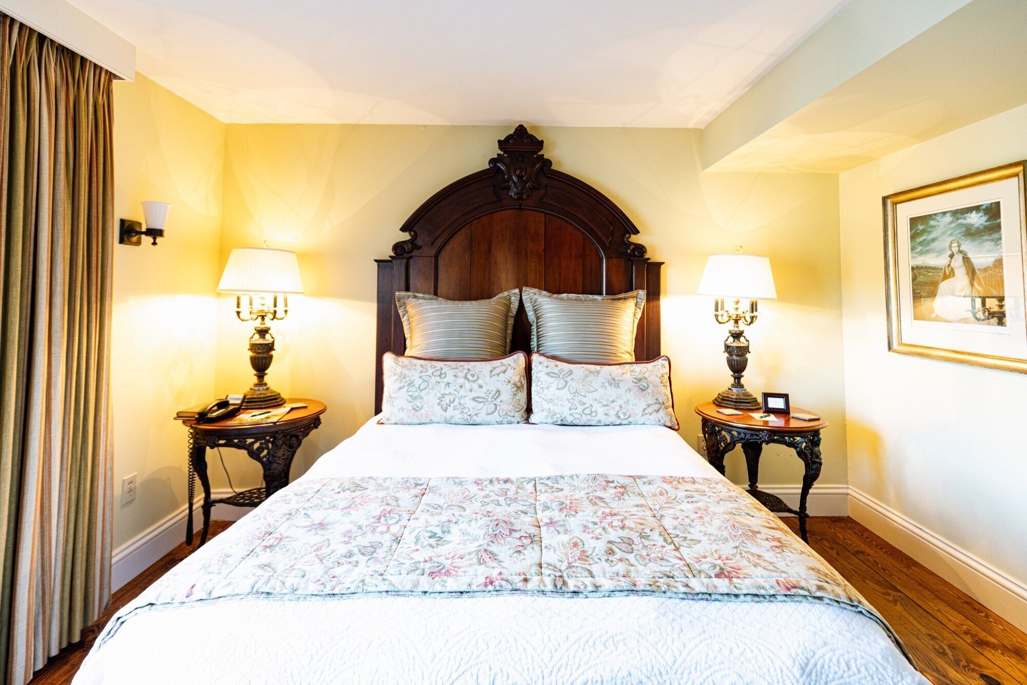 Een hotelkamer met een groot bed, twee nachtkastjes met lampen, een houten hoofdbord, bloemenbedrukte lakens en een ingelijst schilderij aan de muur.