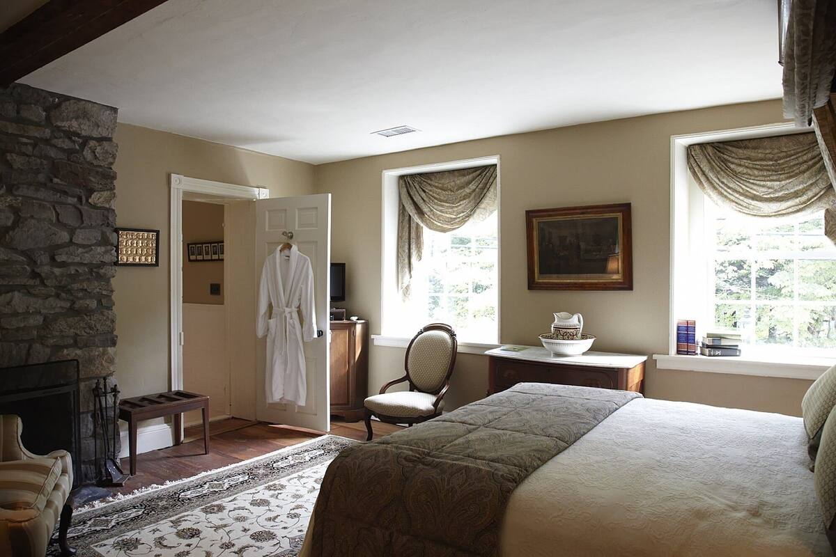 A bedroom with a stone wall, a fireplace, a bed, a chair, a desk with a bowl, and a painting on the wall.